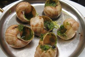 A metal dish with six escargot shells filled with green herb garlic butter, partially eaten, served on a round tray with two gold handles.