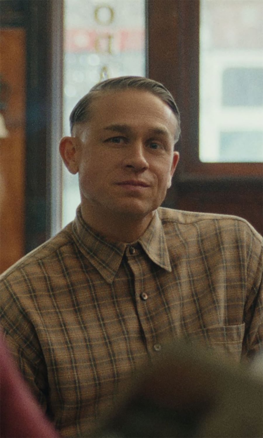 A man with short, neatly combed hair wearing a brown plaid button-up shirt sits indoors, looking slightly to the side with a subtle smile. The background is softly blurred.