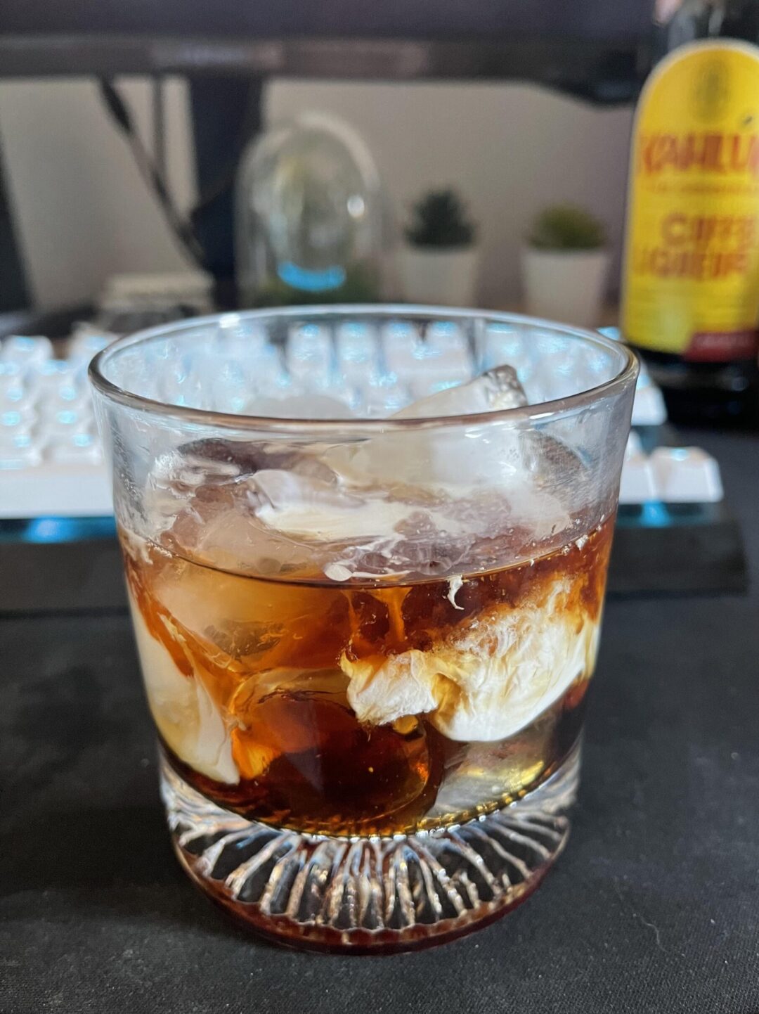 A glass filled with ice, cream, and a dark liqueur, likely a White Russian cocktail, sits on a black surface with a keyboard and a Kahlúa bottle blurred in the background.