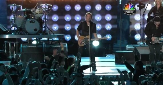 A rock band performs energetically on stage under bright lights, with a cheering crowd in the foreground. The NBC logo appears in the upper right corner of the image.