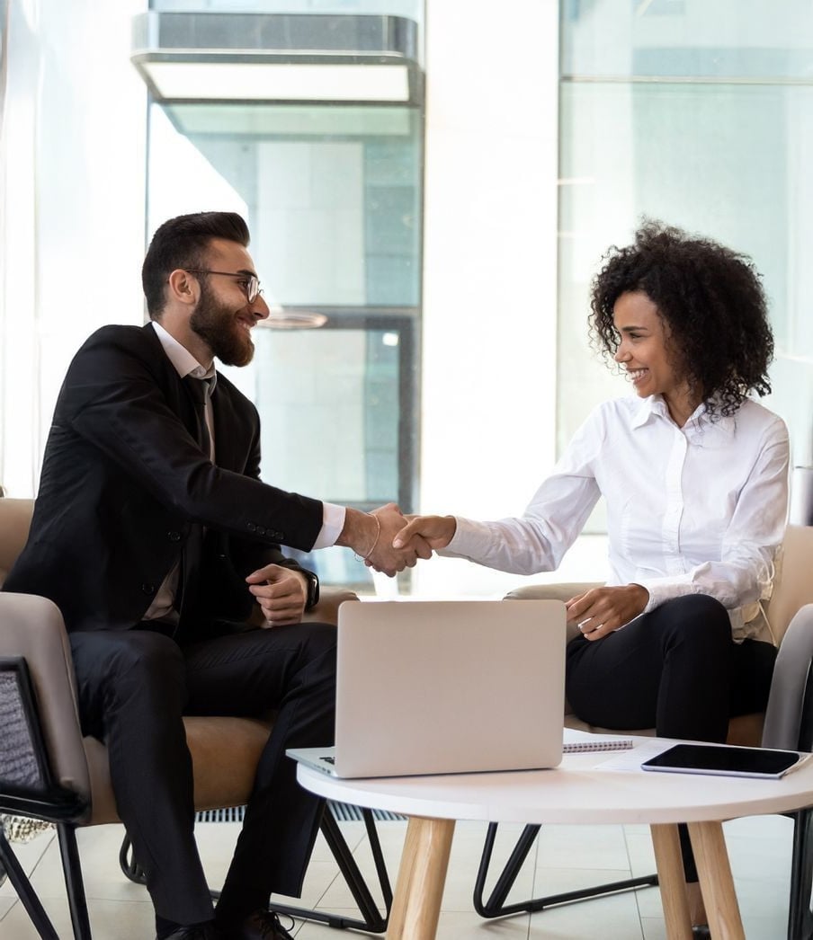 Two business professionals, a man in a suit and a woman in a white shirt, sit in modern chairs and shake hands across a table with a laptop, smiling in a bright office setting.