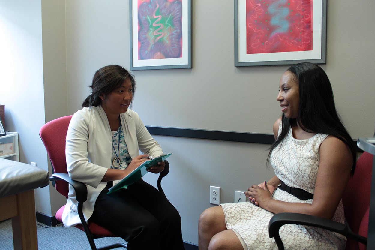 Two women sit facing each other in an office. One, wearing a white coat, holds a clipboard and takes notes; the other, in a white dress, sits with her hands in her lap, listening and smiling. Abstract art hangs on the wall behind them.