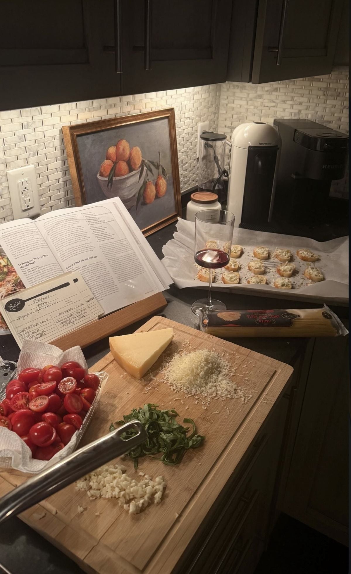 A kitchen counter with chopped tomatoes, garlic, and herbs on a cutting board, a block of cheese, a recipe book, a painting of fruit, a glass of wine, and a tray of prepared food under warm lighting.