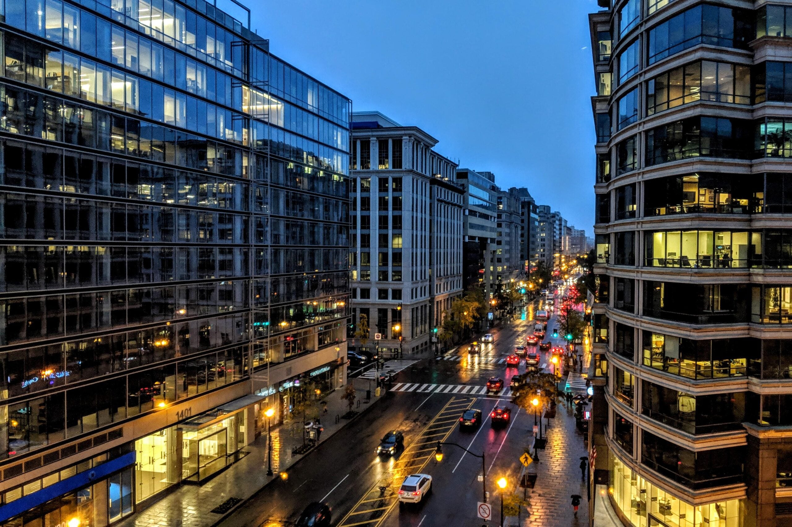 A city street at dusk with tall glass buildings on both sides, wet pavement reflecting lights, and cars driving along the busy road under a cloudy blue sky.