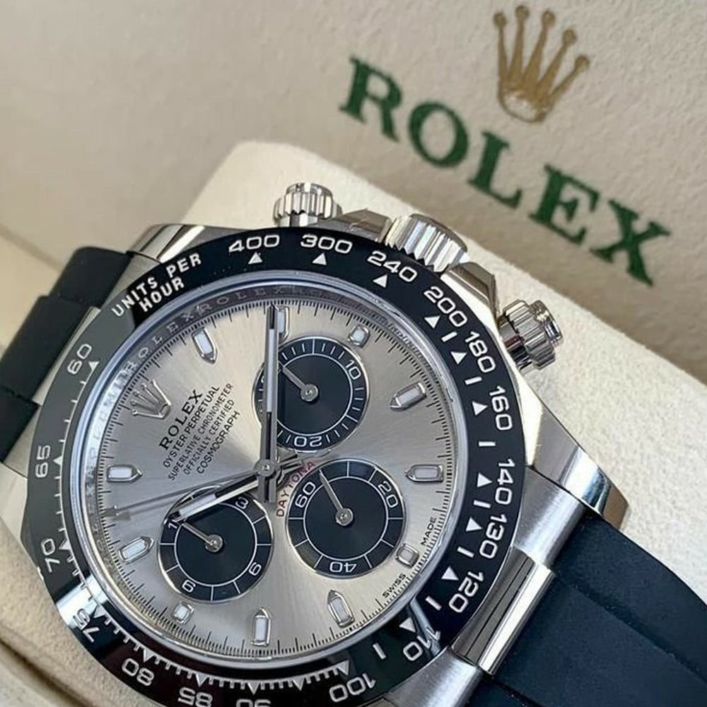 A close-up of a silver Rolex chronograph watch with a black bezel and black rubber strap, displayed in a cream-colored Rolex box with the green Rolex logo and gold crown emblem visible in the background.