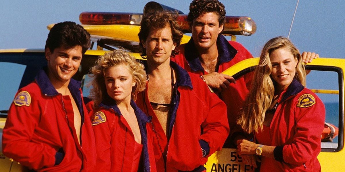 Five people in red lifeguard jackets stand in front of a yellow lifeguard vehicle on a sunny day. They are smiling and posing for the camera, with the ocean and blue sky in the background.