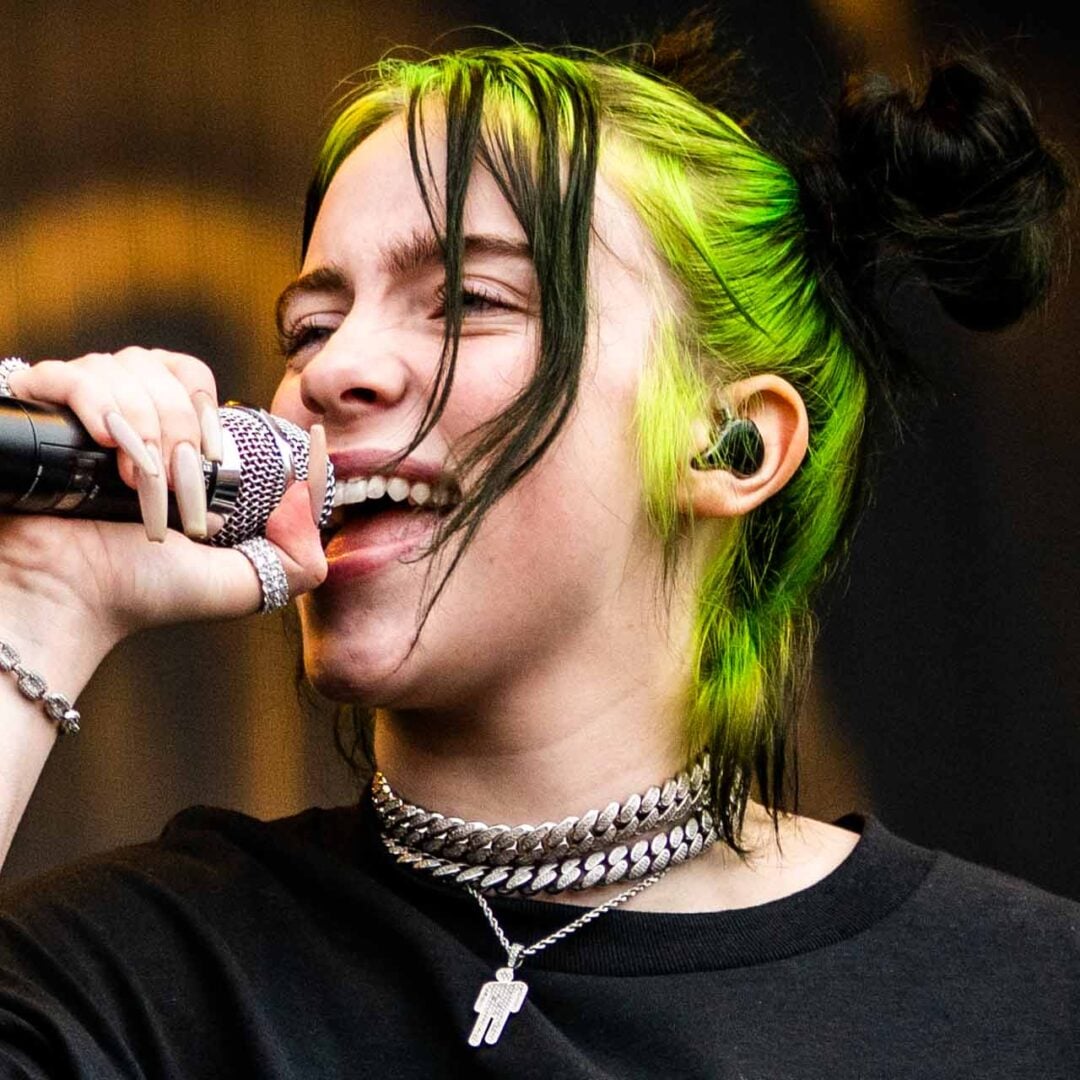 A young person with black and neon green hair sings into a microphone. They wear layered silver chains, a black shirt, and have long, pale nails. Their hair is styled in space buns, and they appear to be performing on stage.