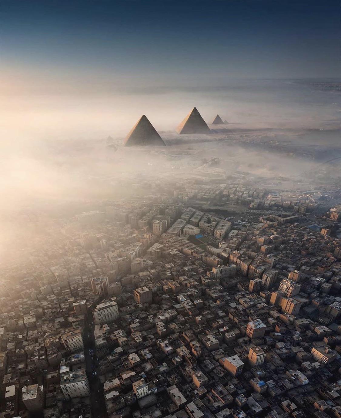 Aerial view of the Pyramids of Giza emerging above thick morning fog, with dense urban buildings of Cairo in the foreground, under a hazy sky.
