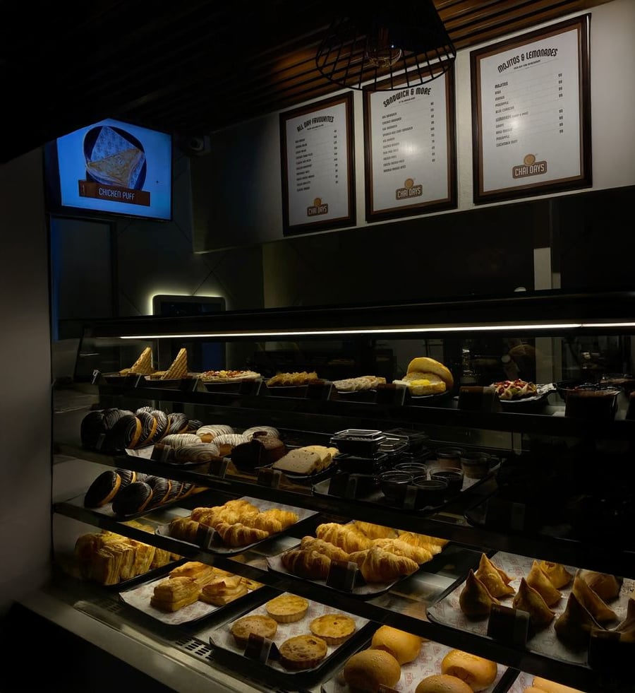 A bakery display case filled with various pastries, bread, and desserts. Above the case are three menu boards and a screen showing “Onion Puff.” The lighting is dim, creating a cozy atmosphere.
