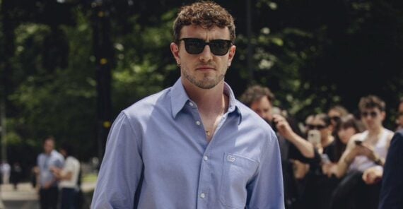 A man in a black suit with an open white shirt sits on a gray chair, one leg crossed over the other, in a stylish room with beige curtains and abstract black-and-white artwork behind him.