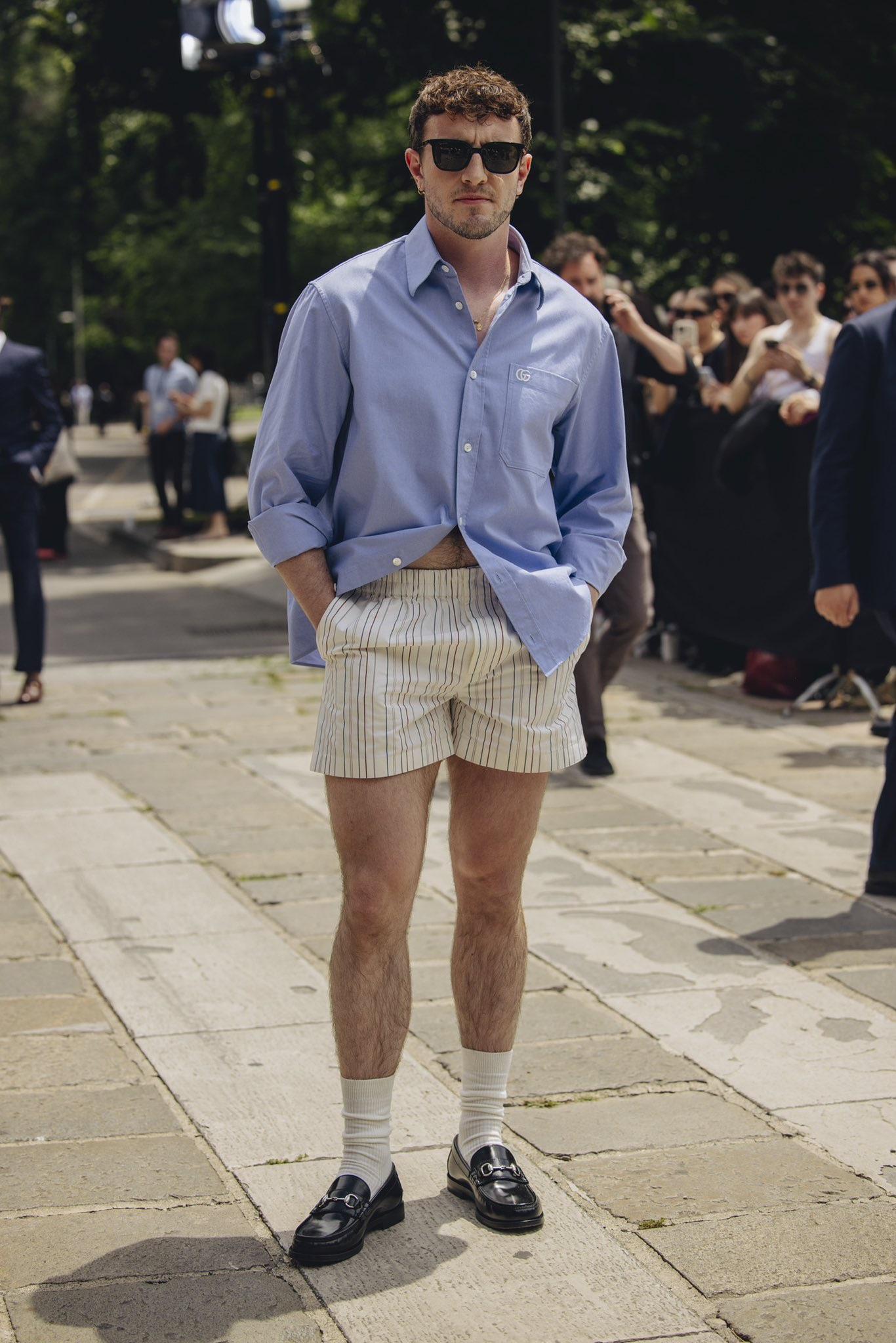 A man wearing sunglasses, a loose blue button-up shirt, striped shorts, white socks, and black loafers stands confidently outdoors on a stone walkway, with people and greenery in the background.