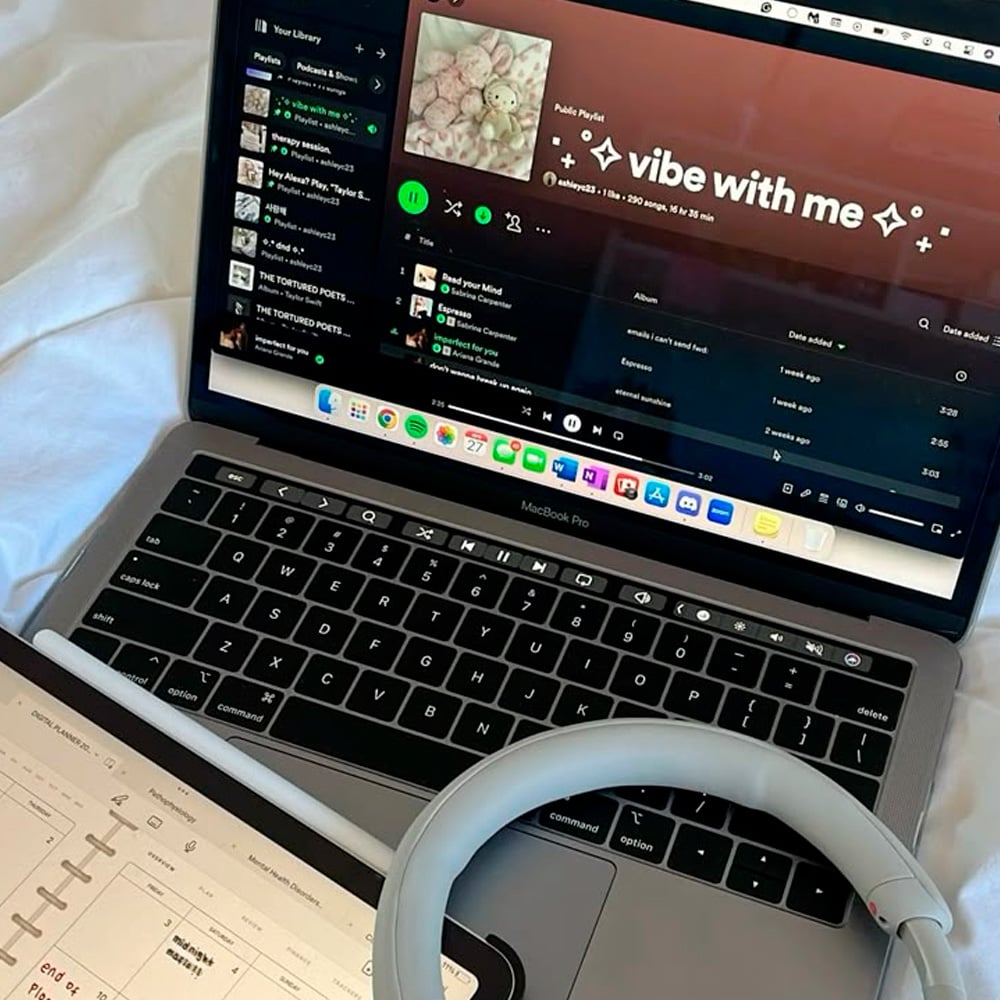 A MacBook Pro displays a Spotify playlist called "vibe with me." Next to it, a tablet shows a digital planner. Overlapping both devices are a white stylus and gray wireless headphones.