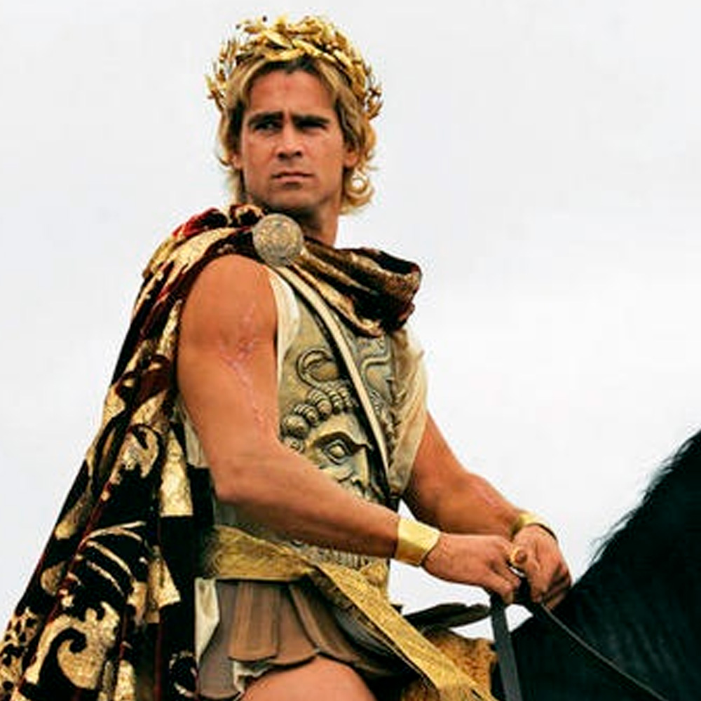 A man dressed as an ancient warrior, wearing a gold laurel crown, ornate armor, and a patterned cape, sits on a black horse against a cloudy sky.