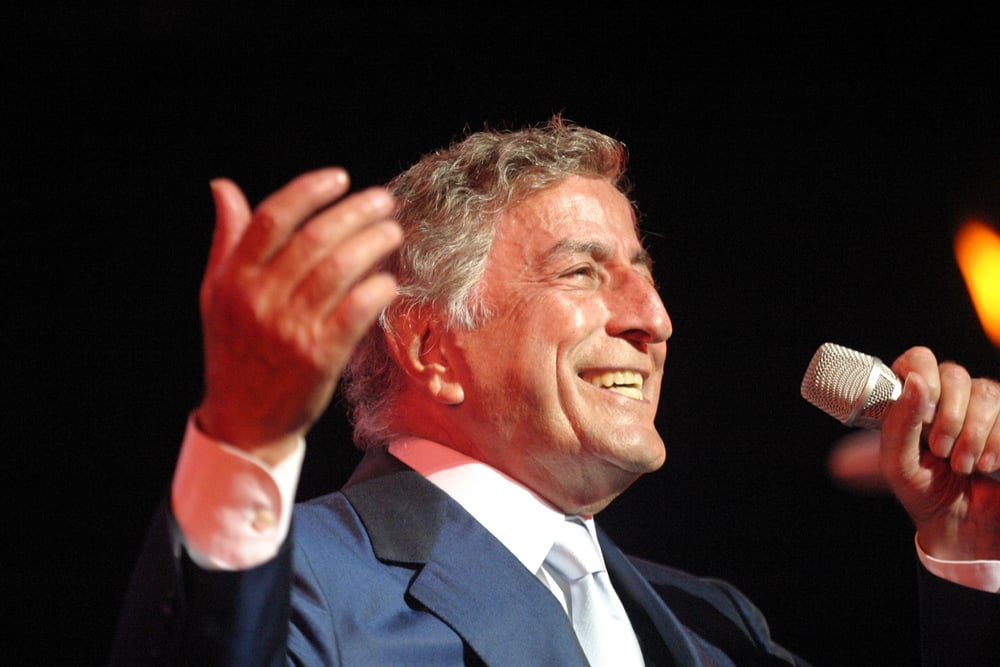 An older man in a blue suit and white shirt smiles while singing into a microphone, holding one hand up expressively. The background is dark, with a small orange light visible.
