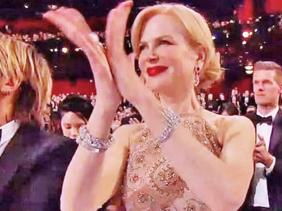 A woman in an elegant, sleeveless dress claps enthusiastically at a formal event, surrounded by other well-dressed attendees in an auditorium with red walls and bright lights.