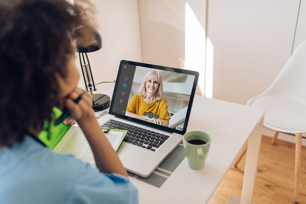 A person sits at a desk with a notebook, looking at a laptop screen displaying a video call with a smiling woman in a yellow shirt. A coffee mug, lamp, and open book are also on the desk.