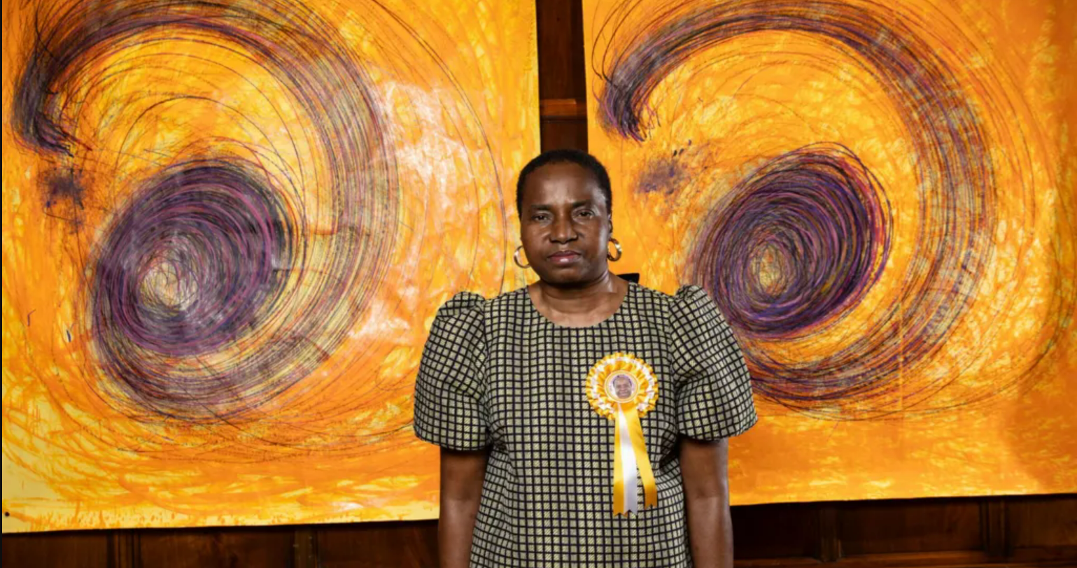A woman wearing a checked dress and a yellow rosette stands in front of two vibrant yellow and purple abstract swirl paintings.