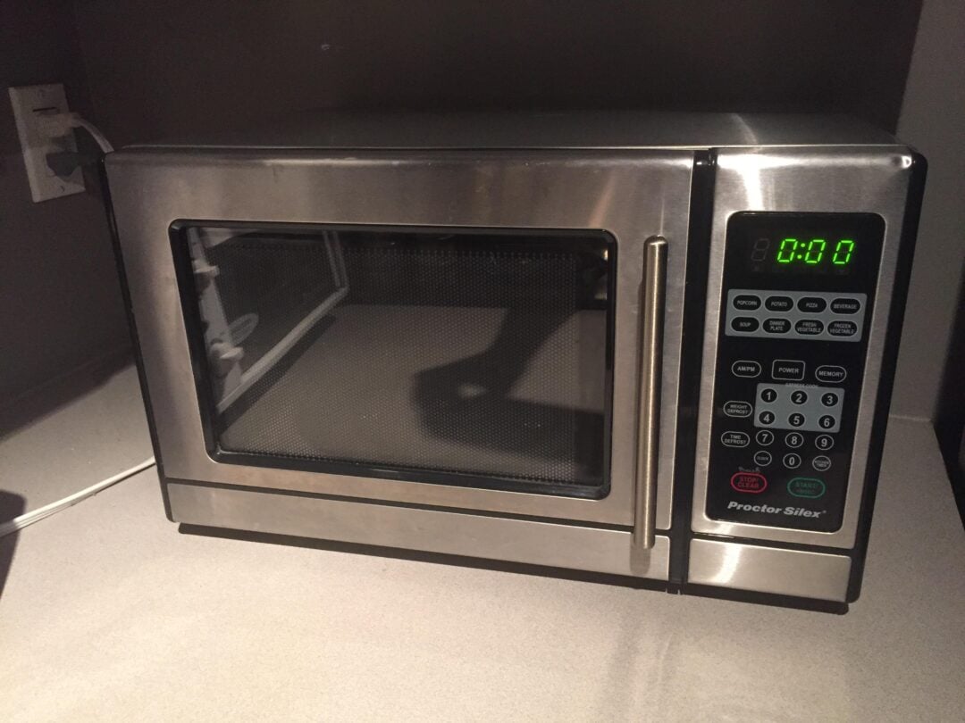 A stainless steel microwave oven sits on a kitchen counter. The display reads "0:00" and the door is closed. The control panel with various buttons is visible on the right side.