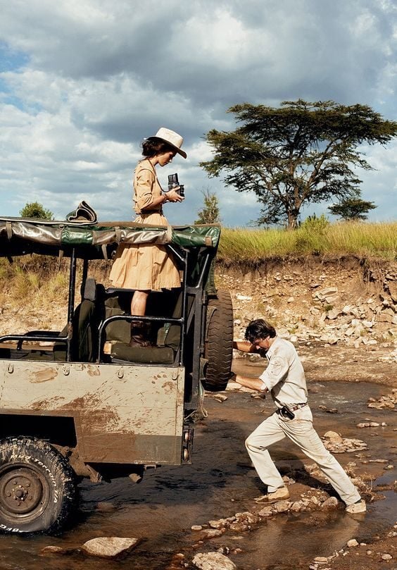 A woman holding a camera stands on the back of a safari jeep while a man in light clothing helps her step down onto rocky ground. They are outdoors near a grassy embankment under a cloudy sky.