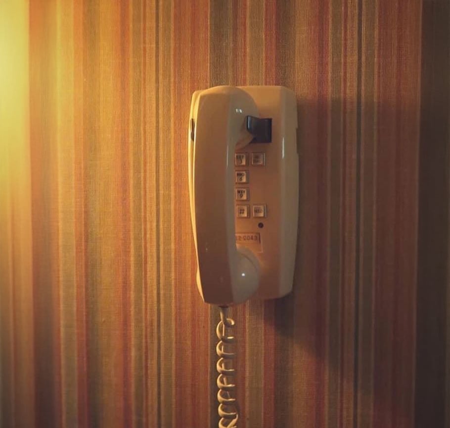 A cream-colored corded wall phone is mounted on a wall with vertical striped wallpaper, bathed in warm, soft light from the left side of the image.