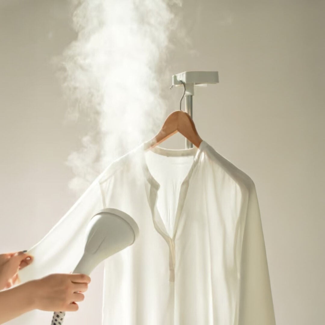 A person uses a handheld steamer to remove wrinkles from a white shirt hanging on a wooden hanger, with visible steam rising.