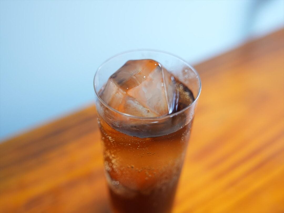 A glass of iced cola with large ice cubes sits on a wooden surface, with visible bubbles rising in the drink. The background is softly blurred.
