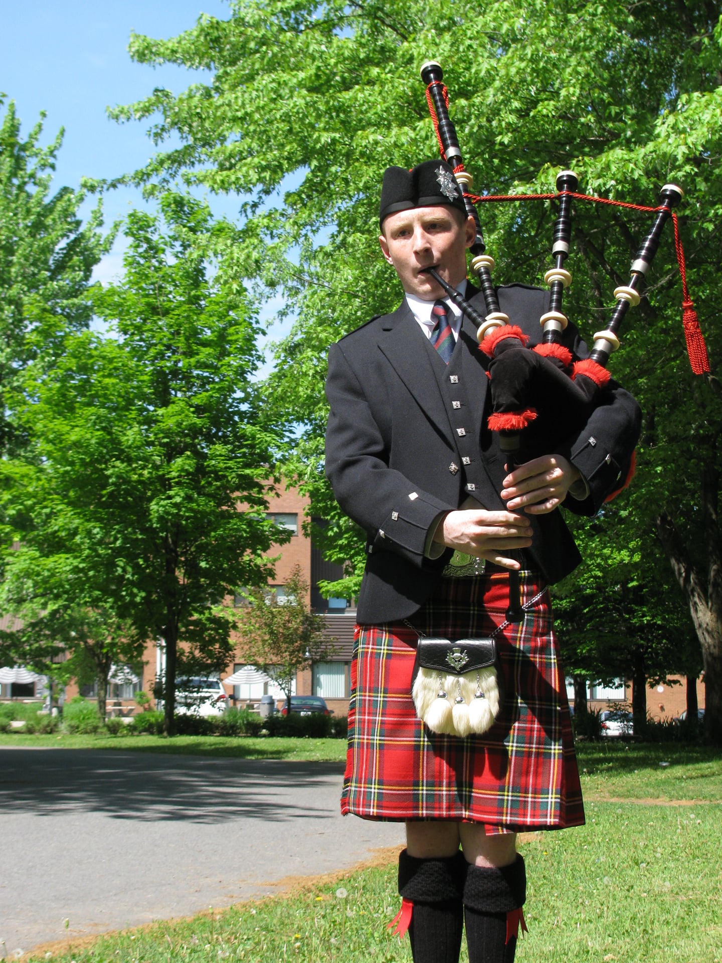 A person wearing traditional Scottish attire, including a kilt and hat, stands outdoors playing the bagpipes in a sunny park with green trees and a building in the background.