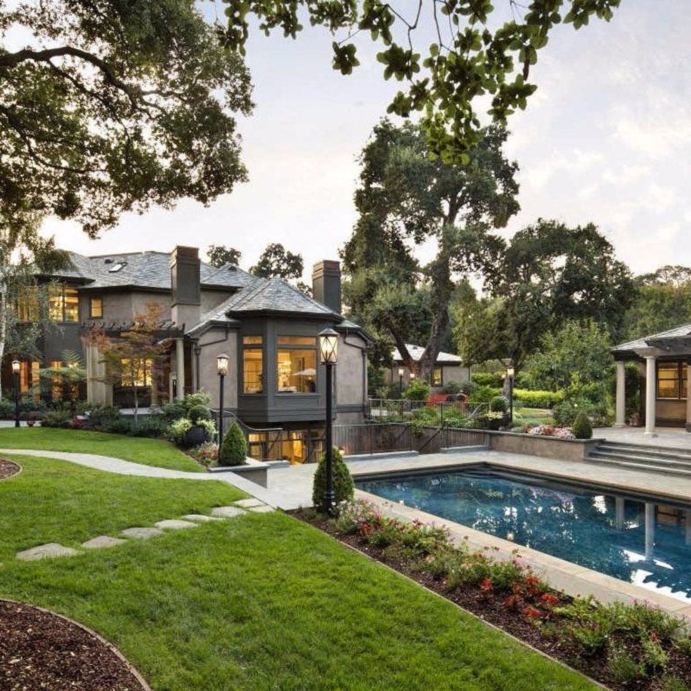 A luxurious two-story house with large windows overlooks a backyard featuring a swimming pool, landscaped garden, green lawn, lampposts, and tall trees surrounding the property.