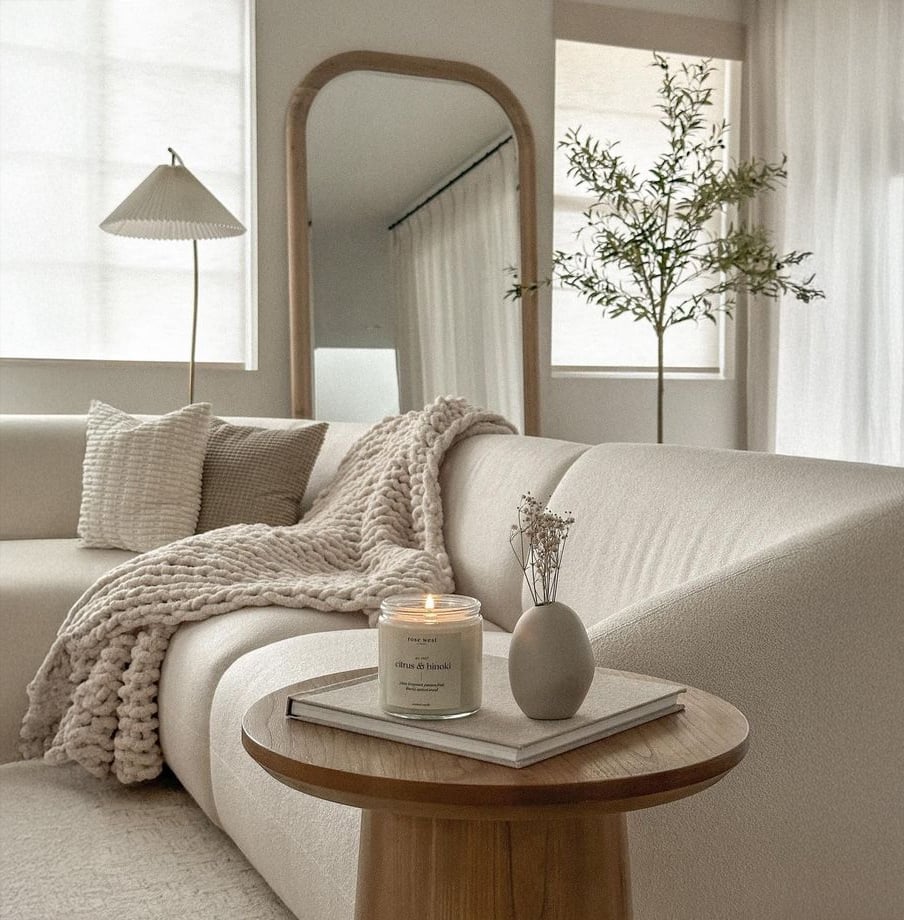 A cozy, minimalist living room with a cream sofa, chunky knit throw, and textured pillow. A wooden table holds a lit candle and small vase. A tall mirror, floor lamp, and potted plant add warmth to the soft, neutral space.