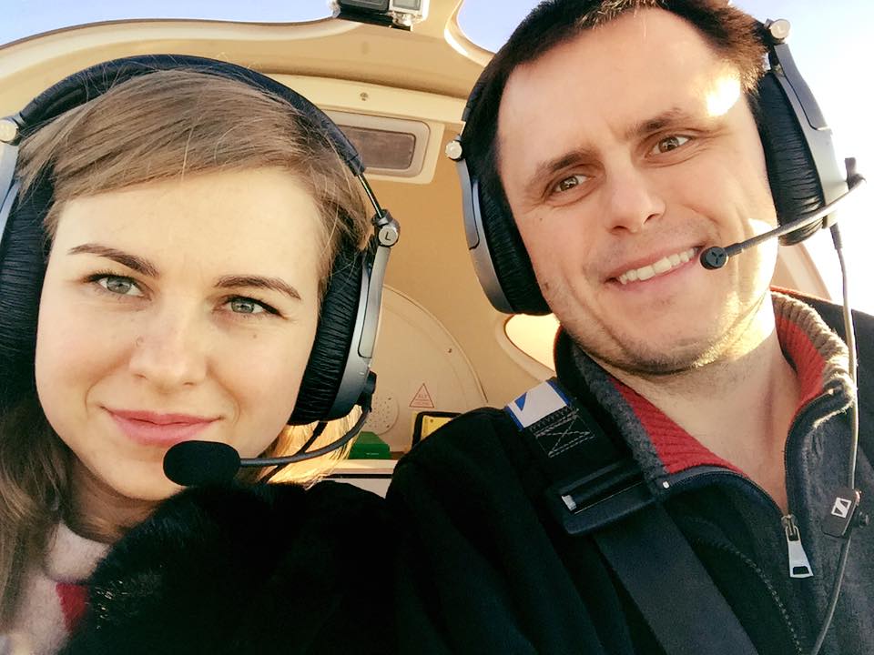A woman and a man wearing headsets with microphones sit side by side inside a small aircraft, smiling at the camera. Sunlight illuminates their faces and the cockpit behind them.