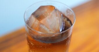 A close-up of a glass filled with a dark, carbonated beverage and a large, clear ice cube, set on a wooden surface.