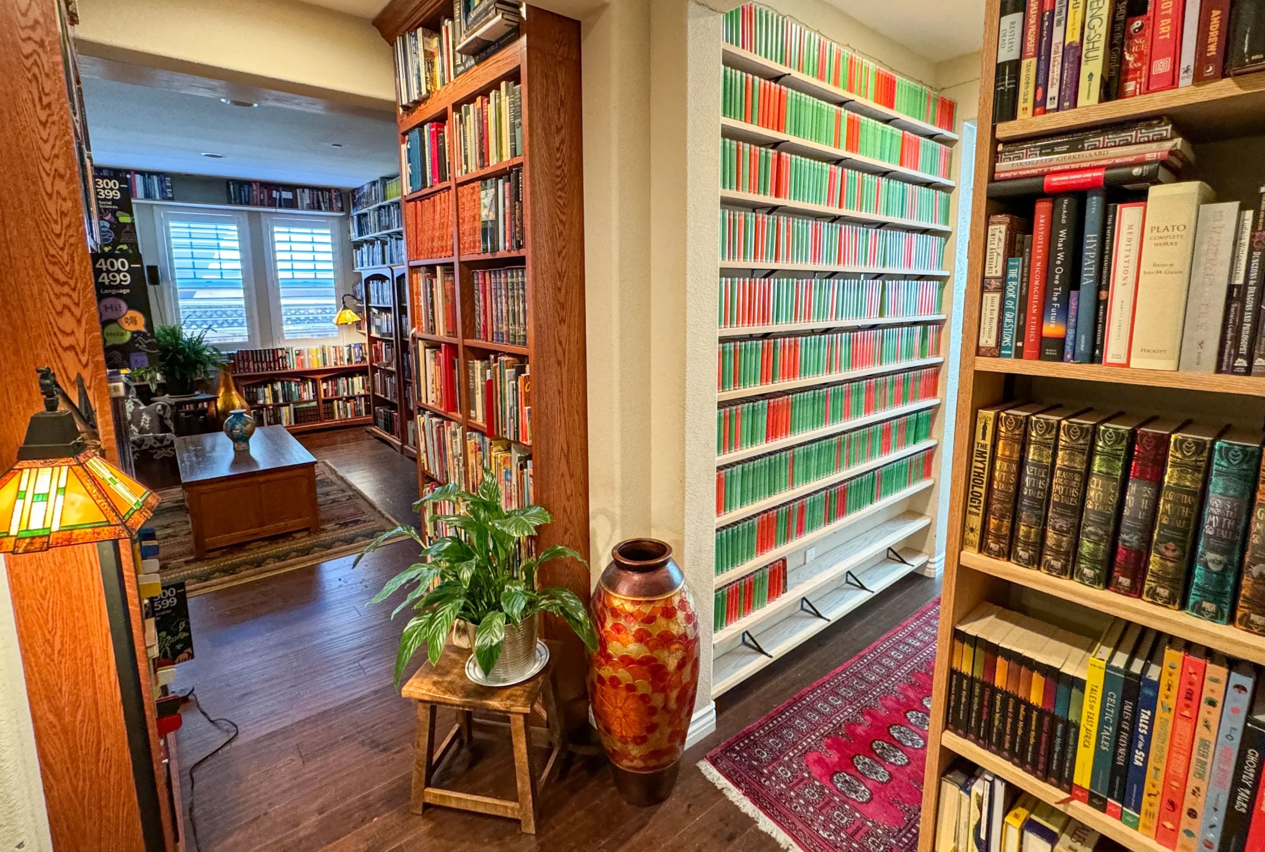 A cozy home library with wooden floors, shelves filled with colorful books, a reading room with a coffee table and chair, a large decorative vase, a potted plant, and a patterned rug by the entrance.
