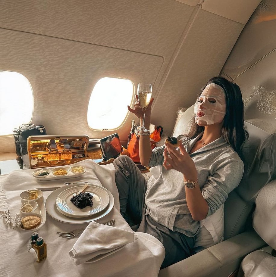 A woman wearing a sheet face mask relaxes in a luxurious airplane seat, holding a glass of champagne and a small dish. A gourmet meal is set on a white tablecloth in front of her, with sunlight streaming through the windows.