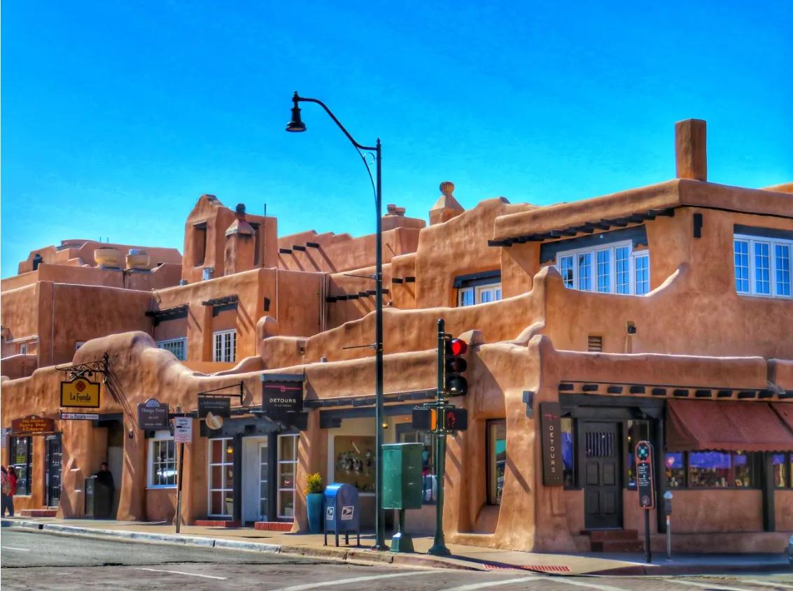 Adobe-style southwestern buildings with rounded edges and tan stucco façades sit on a street corner under a bright blue sky. Shops and signs are visible at street level, with a traffic light and streetlamp in front.