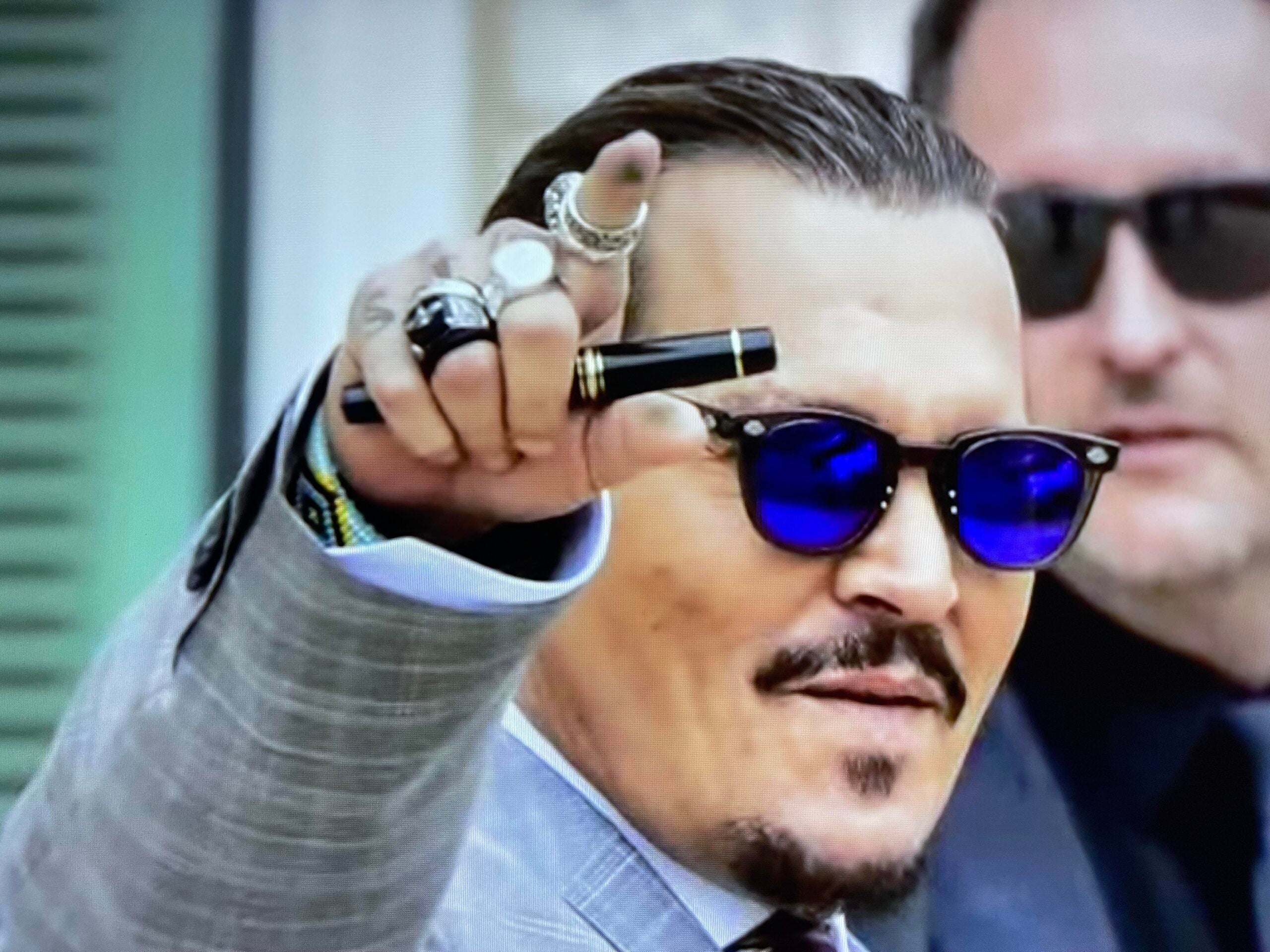 A man wearing a gray suit, blue-tinted sunglasses, and several rings points forward while holding a pen. Another person in sunglasses stands behind him.