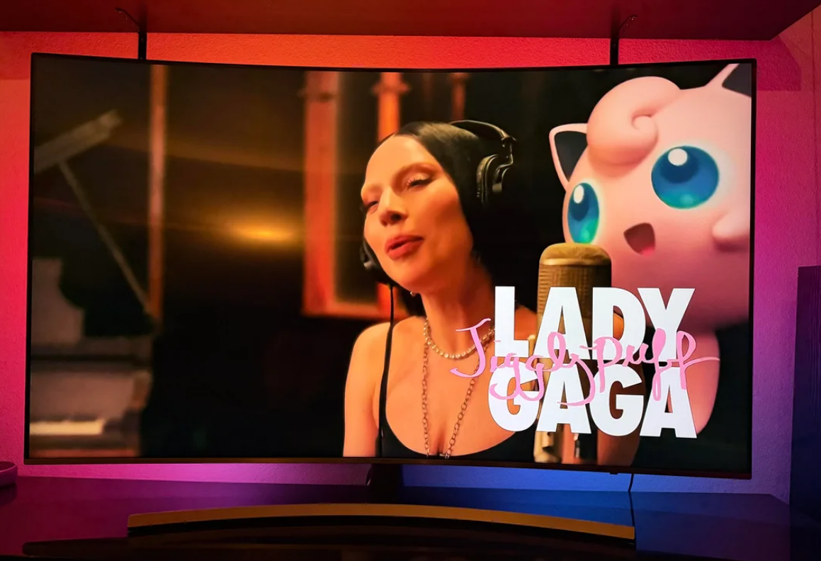 A woman in a recording studio sings into a microphone, with the words "Lady Gaga" overlaid and a large animated Jigglypuff character beside her. The scene is lit with colorful, warm lighting.