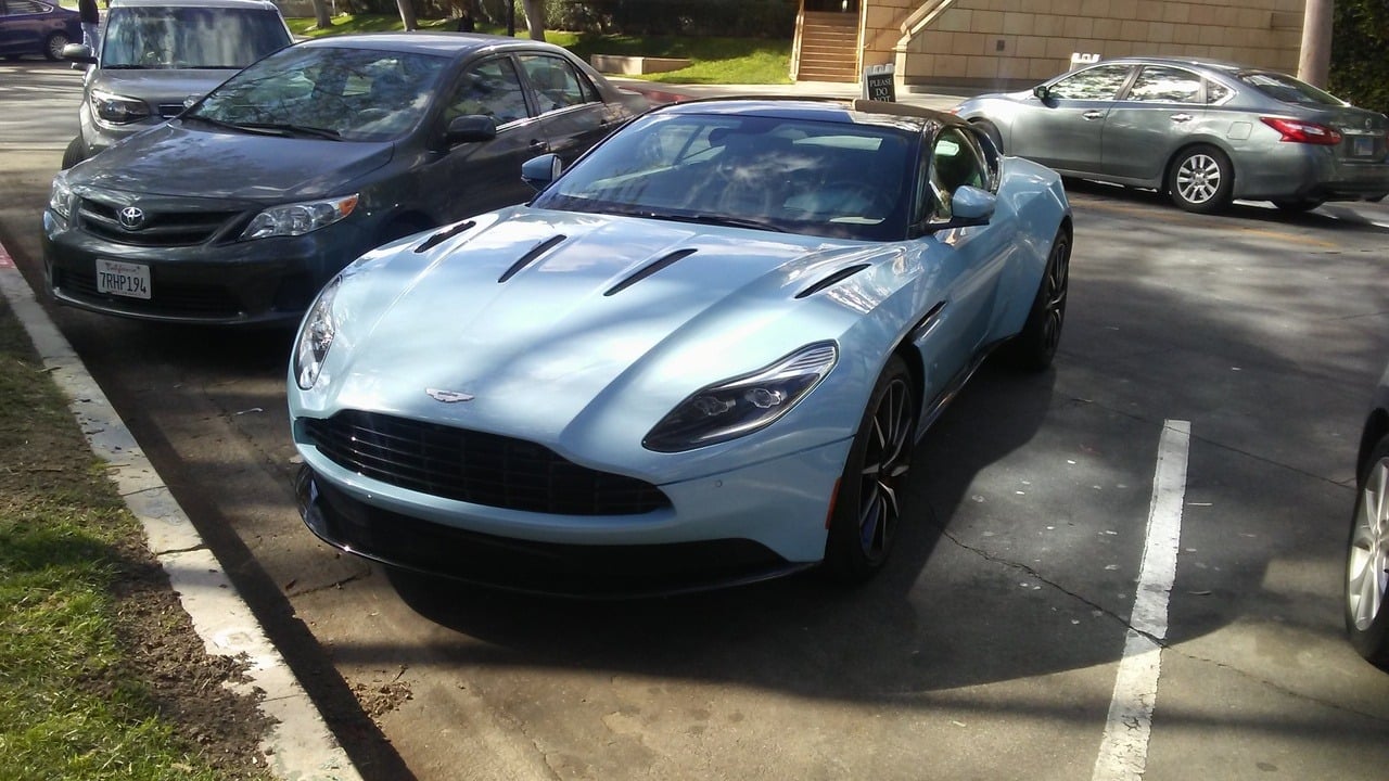 A light blue Aston Martin sports car is parked in a parking lot between two gray sedans on a sunny day.