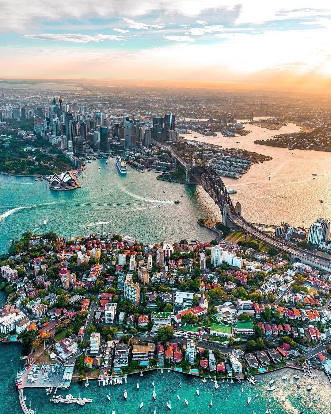 Aerial view of Sydney at sunset, featuring the Sydney Opera House, Sydney Harbour Bridge, city skyline, and waterfront neighborhoods surrounded by boats and calm water.