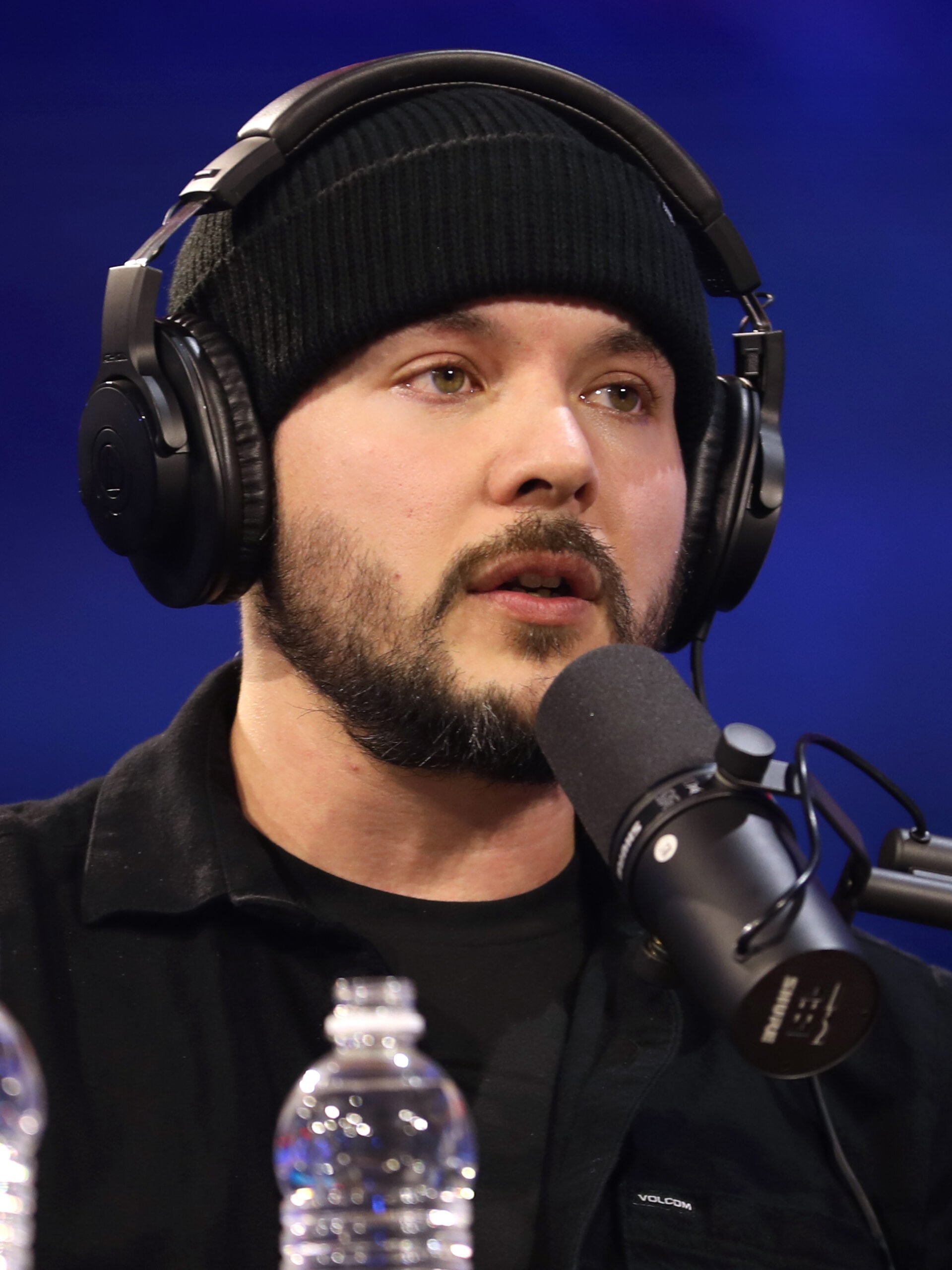 A man with a beard wearing a black beanie and black headphones speaks into a microphone in a podcast studio. A water bottle and blurred background are visible in the foreground.