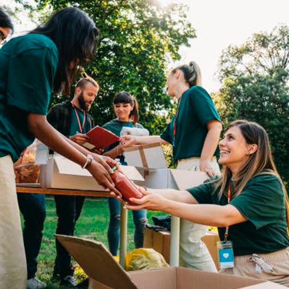 A group of volunteers wearing green shirts are sorting and packing donated goods into boxes outdoors on a sunny day, smiling and working together.