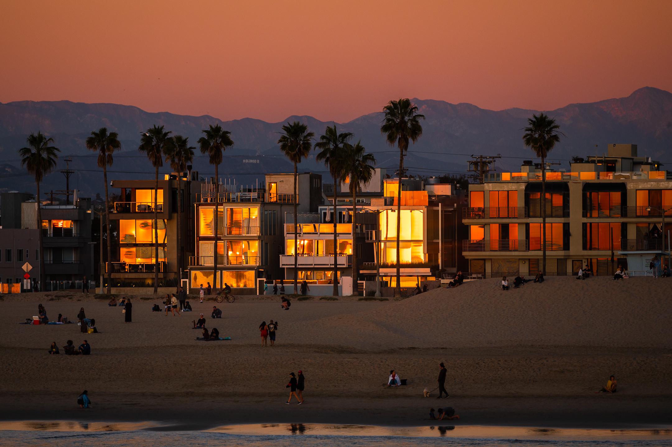People relax on a sandy beach at sunset, with modern buildings and palm trees in the background reflecting the orange glow of the setting sun. Mountains rise in the distance under a clear sky.