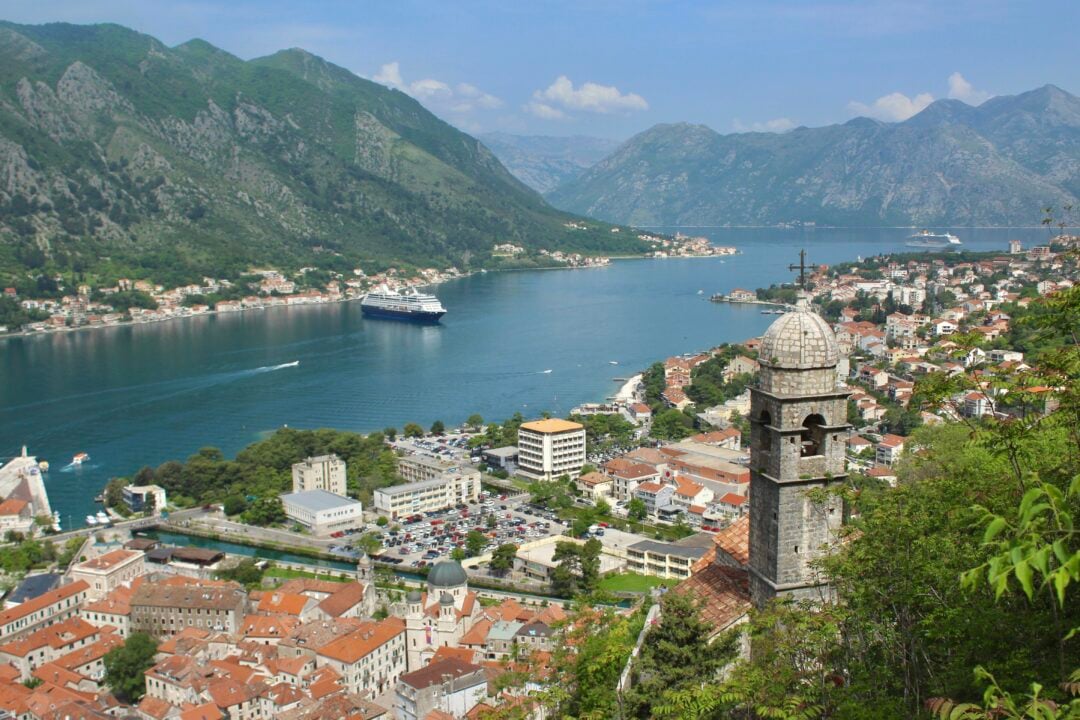 A scenic view of Kotor Bay in Montenegro, showing a coastal town with red-roofed buildings, a stone church with a bell tower, lush green mountains, and cruise ships docked in the blue bay under a partly cloudy sky.