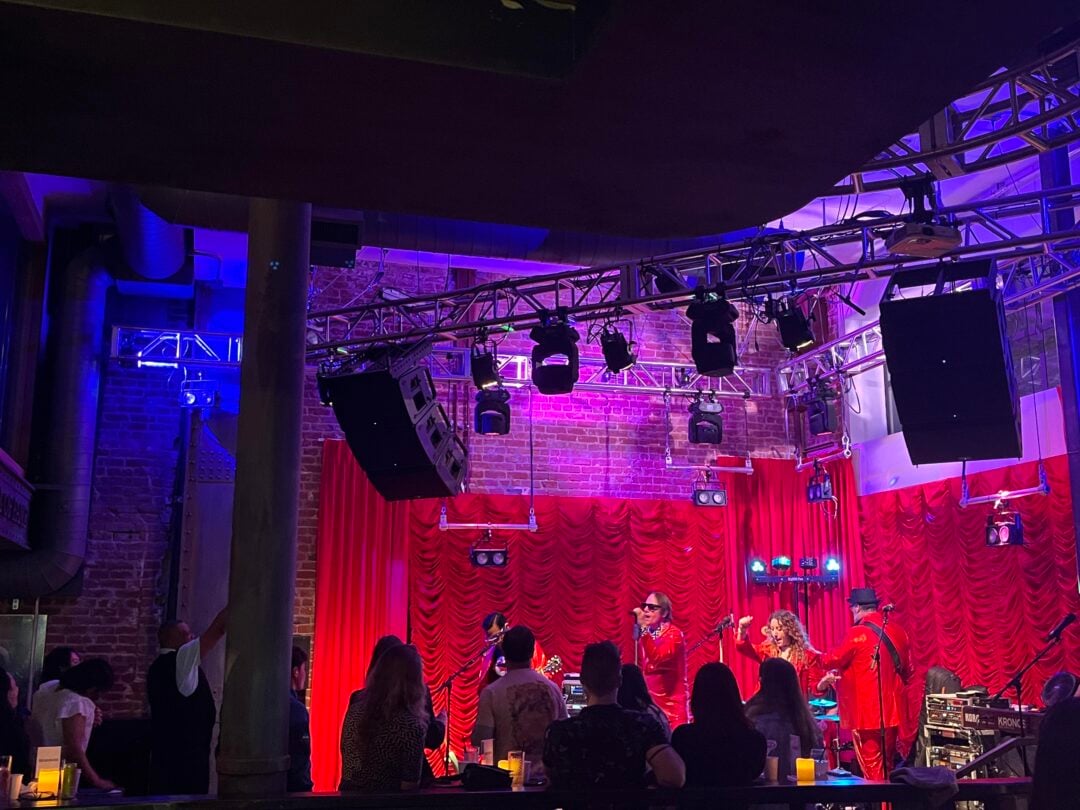 A band performs on a stage with red curtains and colorful lighting in a brick-walled venue, while an audience watches from tables with glowing candles.