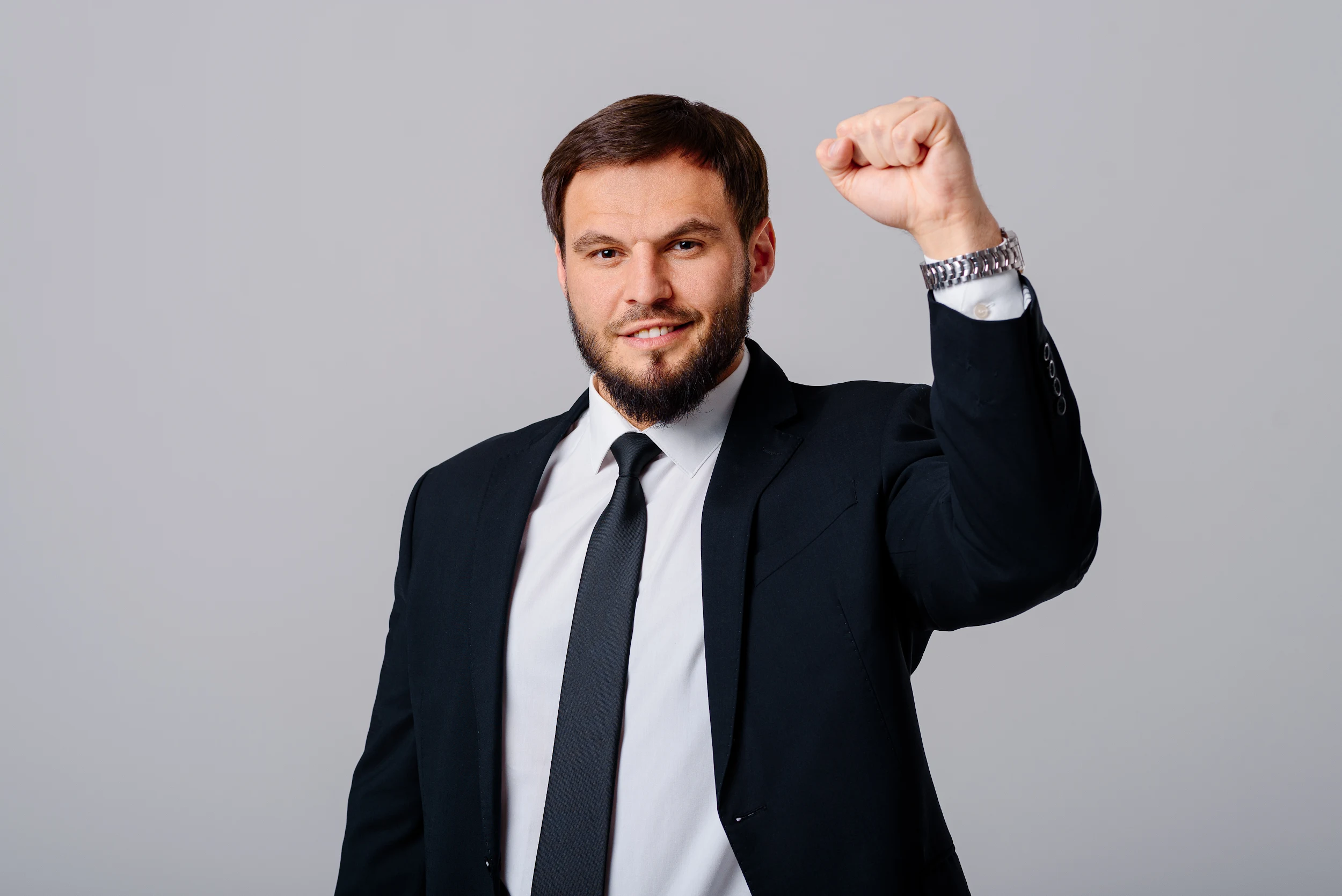 A man in a black suit and tie stands against a plain gray background, raising his right fist in the air in a gesture of strength or celebration, and smiling confidently at the camera.