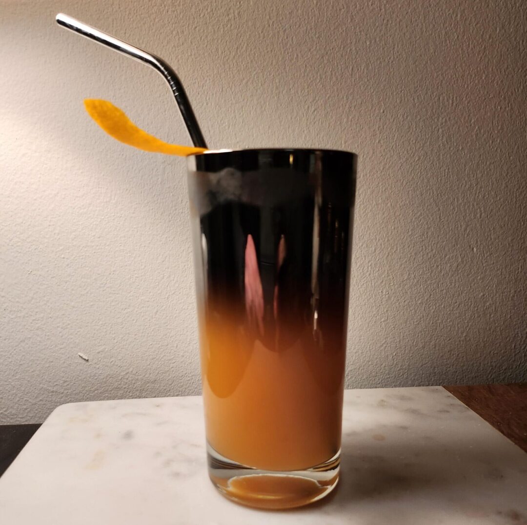 A tall glass filled with a layered orange and brown drink sits on a marble surface. The glass has a metal straw and an orange peel garnish, with a soft light casting a gentle shadow on the wall behind.