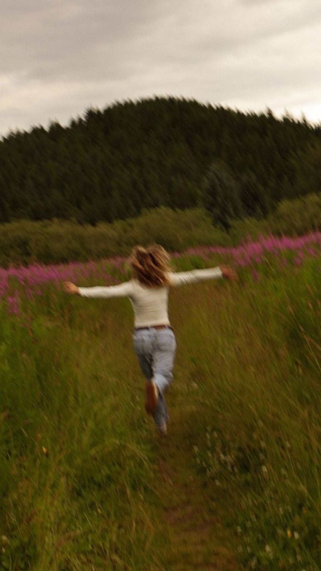 A person with long hair runs joyfully down a grassy path surrounded by tall green grass and wildflowers, with a forested hill in the background under a cloudy sky.