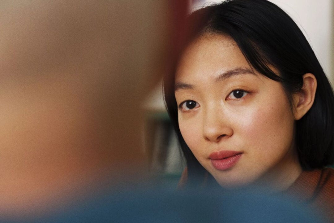 A woman with straight black hair and natural makeup looks attentively at someone whose blurred head is in the foreground, creating a sense of intimate conversation.