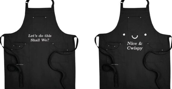 Two black aprons are shown side by side. The left apron says, &ldquo;Let&rsquo;s do this Shall We?&rdquo; and the right apron says, &ldquo;Nice & Cwispy&rdquo; with a small smiley face above the text.