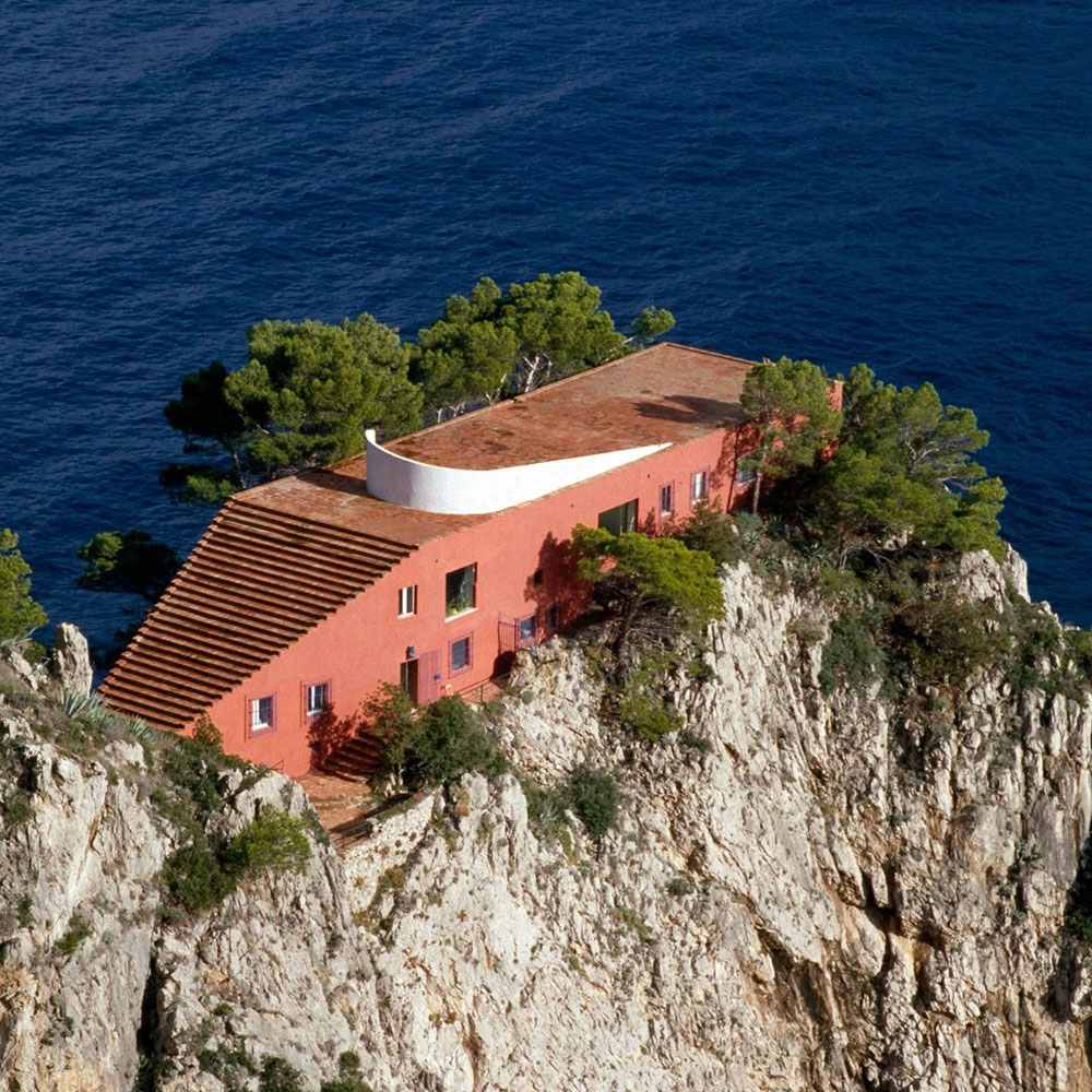 A red modern house with a sloped roof sits atop a rocky cliff surrounded by trees, overlooking a deep blue ocean.
