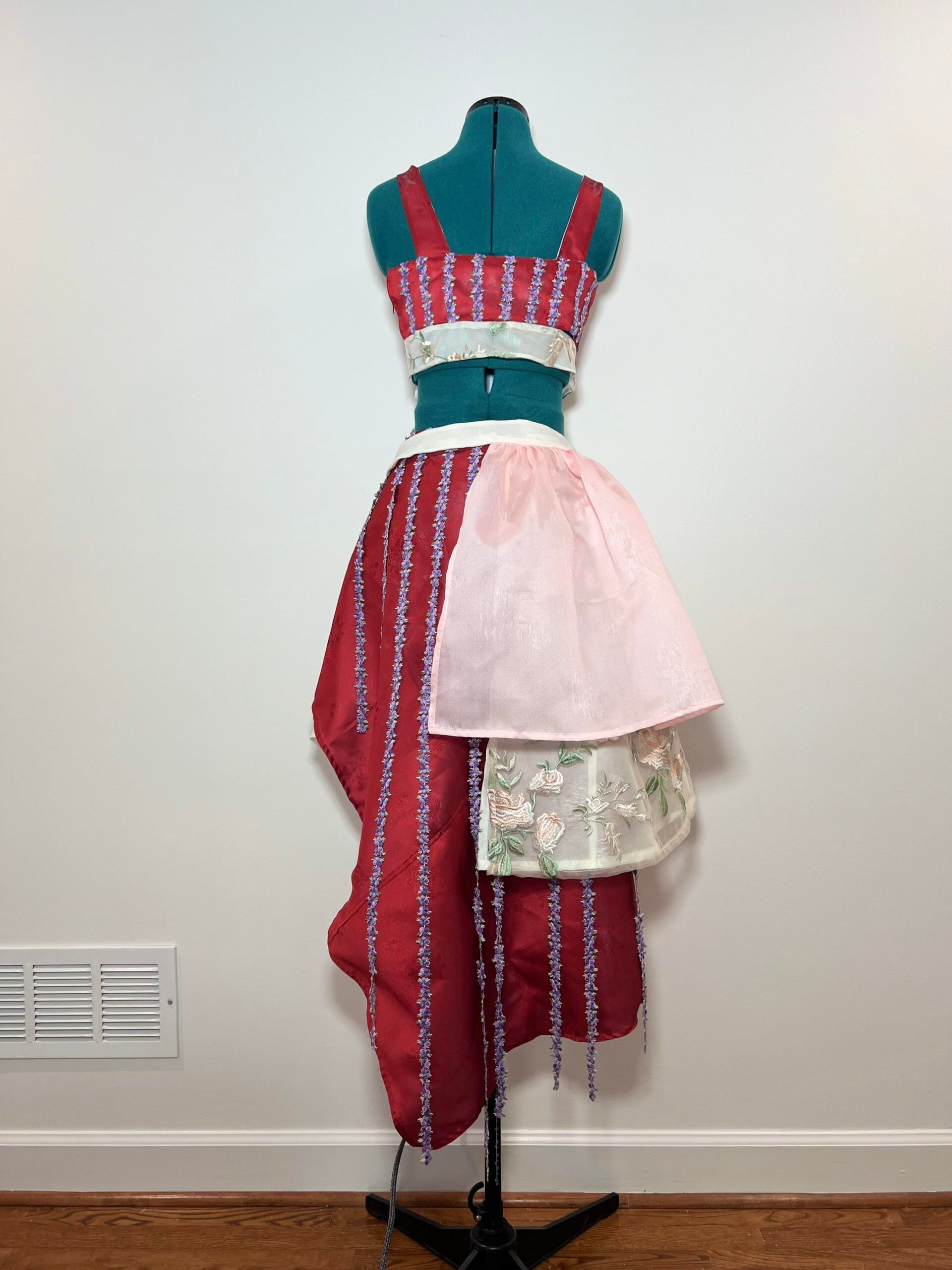 A dress form displays a unique outfit featuring a red and lavender striped crop top and an asymmetrical skirt with red panels, a sheer pink overlay, and a white floral embroidered section, set against a plain wall.